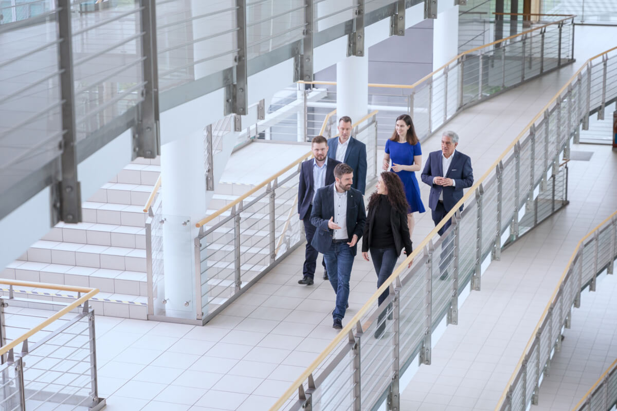 Six business people are walking down a corridor in a modern office building and talking.