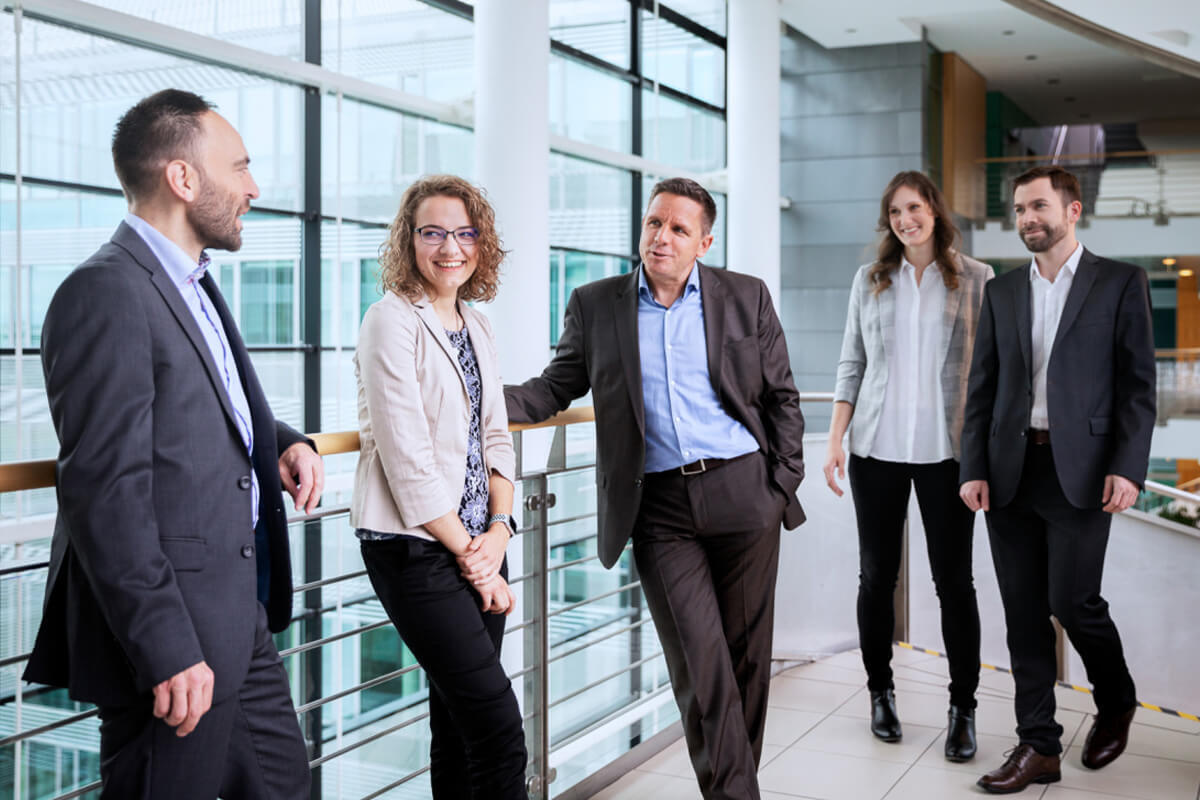 A group of five business people in business attire stand together in a modern office atrium and have a conversation.