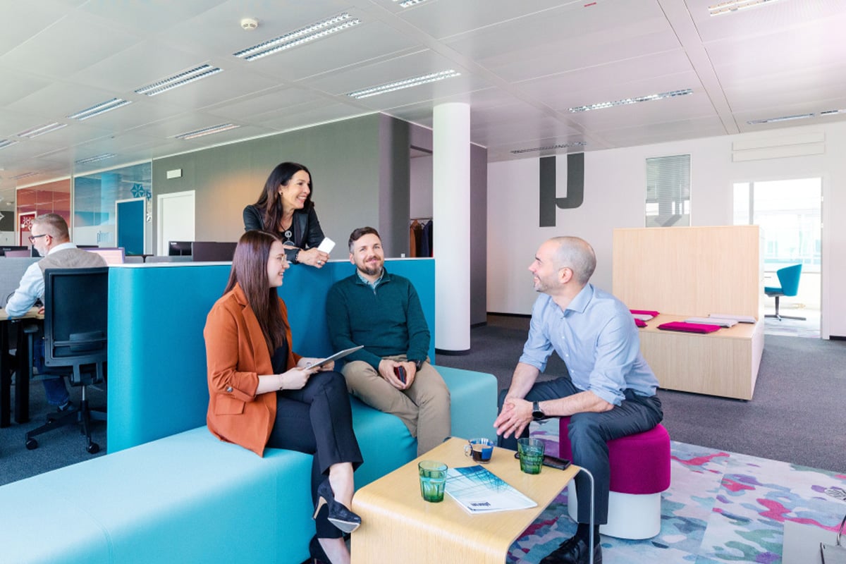 A group of experts discuss in a modern office space with colorful seating and a collaborative atmosphere.