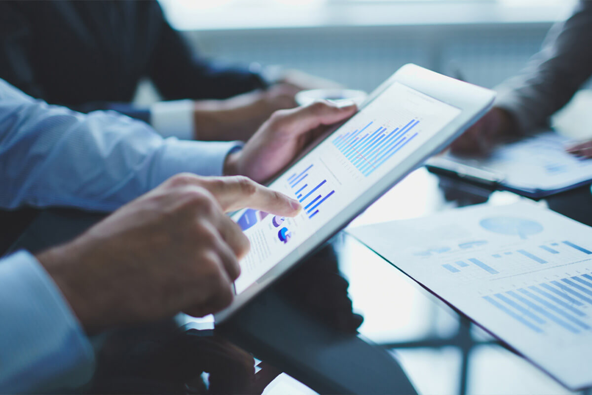 Close-up of hands interacting with a tablet displaying data graphics; reports on a conference table can be seen in the background.