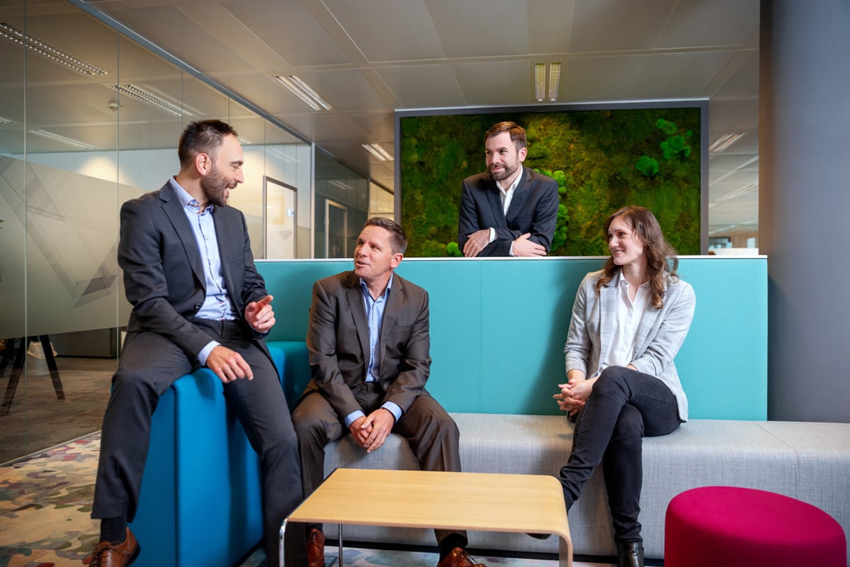 Eine Gruppe von vier Personen in Businesskleidung sitzt in einer modernen Bürolounge und spricht miteinander.