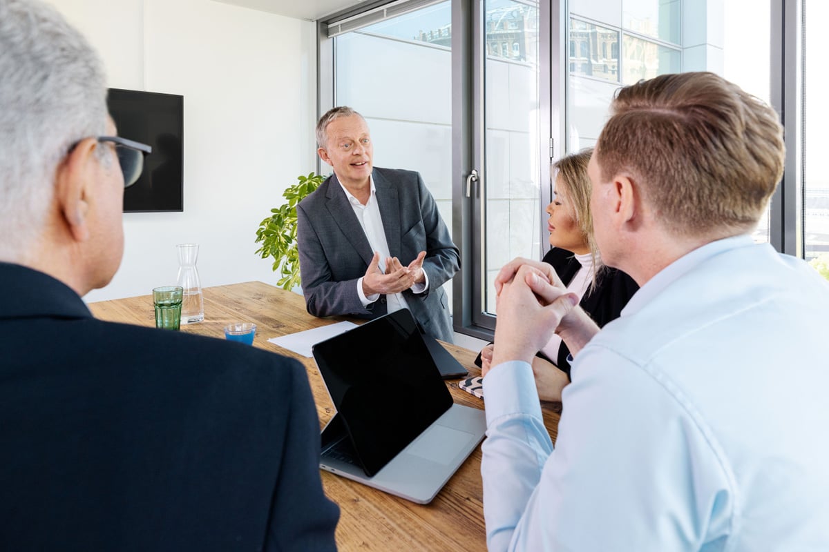 A group of experts are discussing at a wooden table in a bright, modern office space. A tablet is on the table.