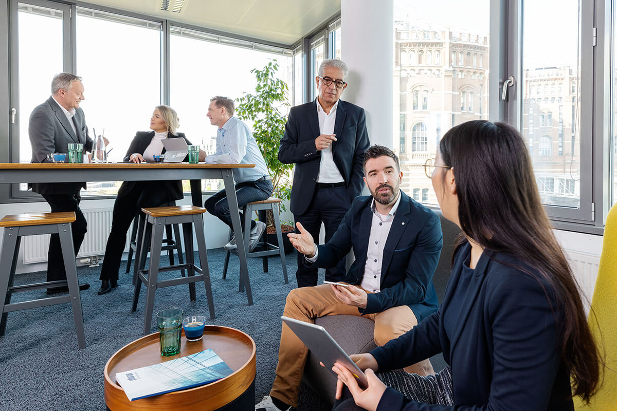 A professional meeting takes place in a bright office space where participants discuss at a high table while others sit nearby.