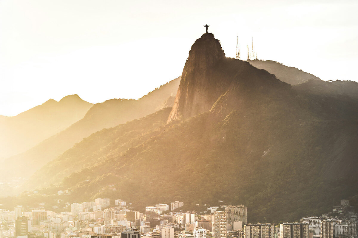 The iconic statue of Christ the Redeemer on Corcovado Mountain overlooks Rio de Janeiro at sunset, surrounded by buildings and mountains.