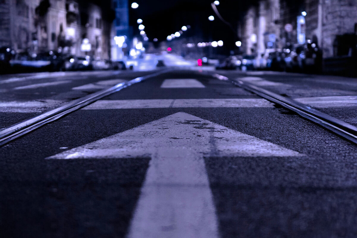 A dimly lit street at night with a striking white directional arrow, streetcar tracks and blurred city lights in the background.