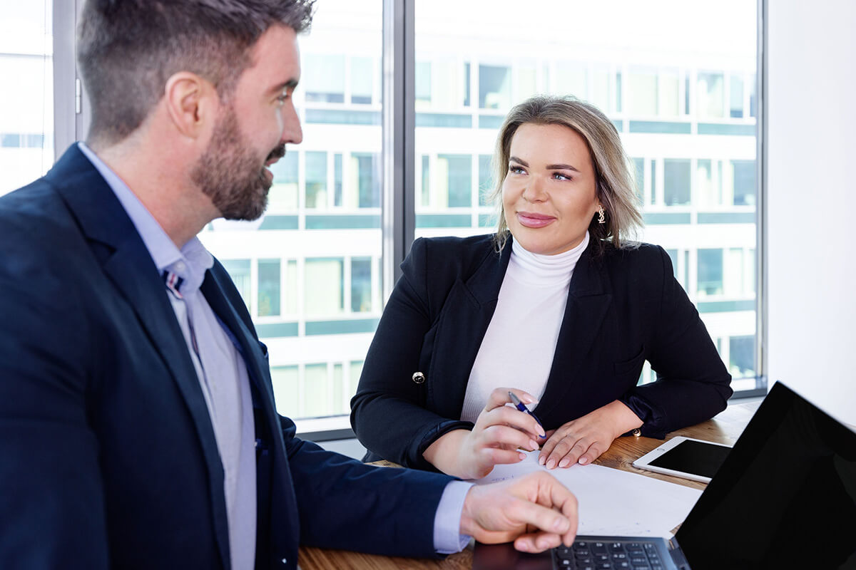 A meeting between two people in business attire discussing notes and using a laptop in a modern office environment.