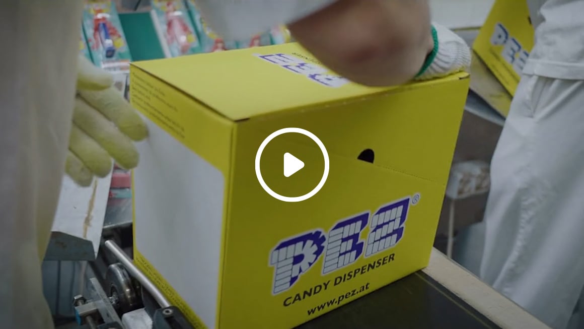 Video still: A worker wearing gloves is handling a yellow PEZ receipt can on a conveyor belt in a production plant.