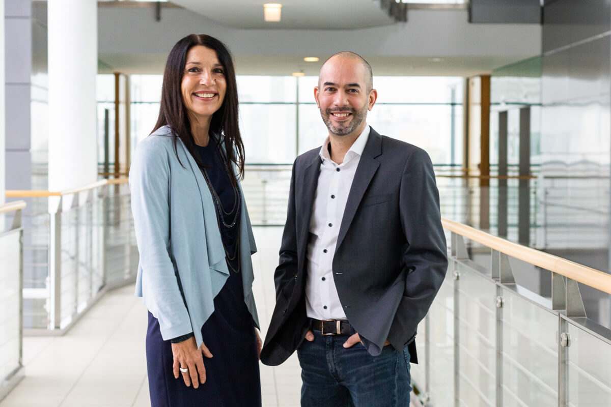 Porträtbild mit Sandra Spies und Michael Perfler, die nebeneinander in einem modernen, hellen Büroflur stehen.