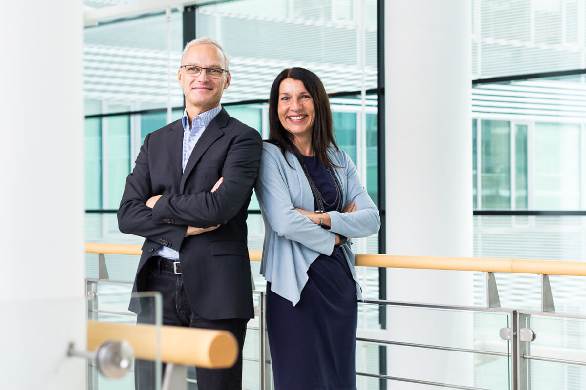Porträtbild mit Sandra Spies und Friedrich Lammer, die nebeneinander in einem modernen, hellen Büroflur stehen.