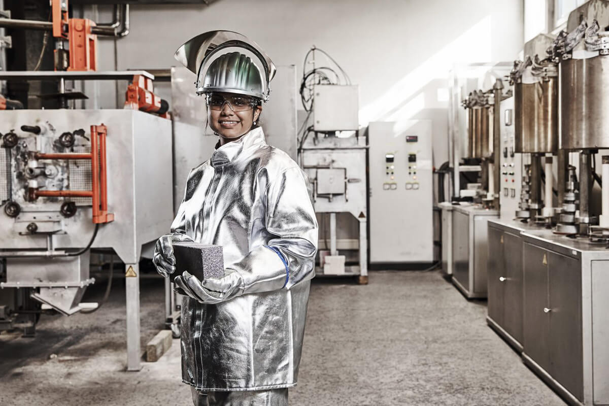 Eine Arbeiterin in einem silberglänzenden Schutzanzug hält ein dunkles rechteckiges Objekt in den Händen und steht in einer modernen Industrieanlage mit Maschinen im Hintergrund.