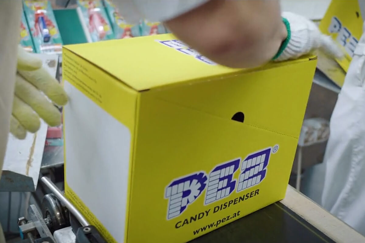 A worker wearing gloves is handling a yellow PEZ receipt can on a conveyor belt in a production plant.
