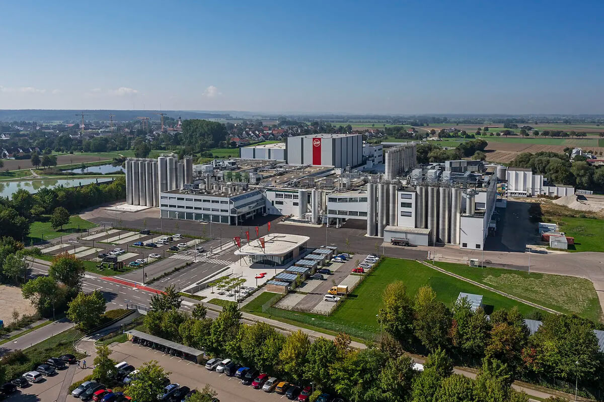 Luftaufnahme des ZOTT-Werks in Mertingen – eine Industrieanlage mit Silos, Parkplätzen und Grünflächen unter strahlend blauem Himmel.